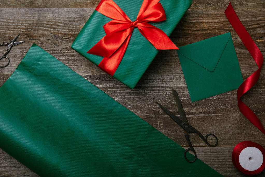 top view of wrapped christmas gift, scissors, blank envelope and green wrapping paper on wooden background - Photo, Image