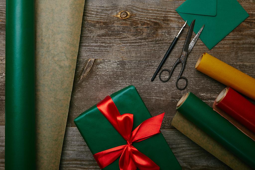 flat lay with wrapped christmas present with ribbon on wooden surface with wrapping papers, envelope and scissors - Photo, Image