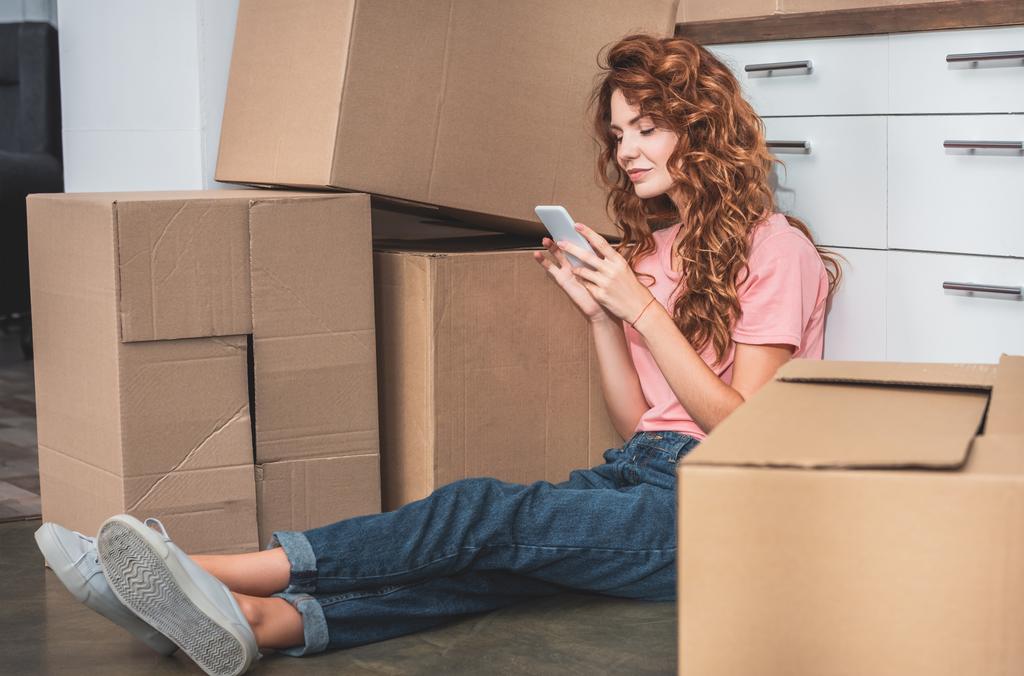 привлекательная женщина с вьющимися волосами сидит на полу рядом с картонными коробками и использует смартфон в новом доме
 - Фото, изображение