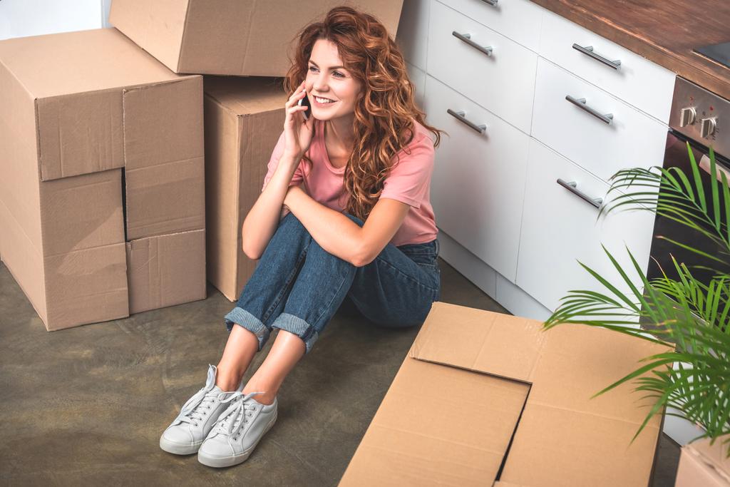 lächelnde attraktive Frau mit lockigem Haar sitzt auf dem Boden neben Pappkartons und unterhält sich im neuen Zuhause per Smartphone - Foto, Bild