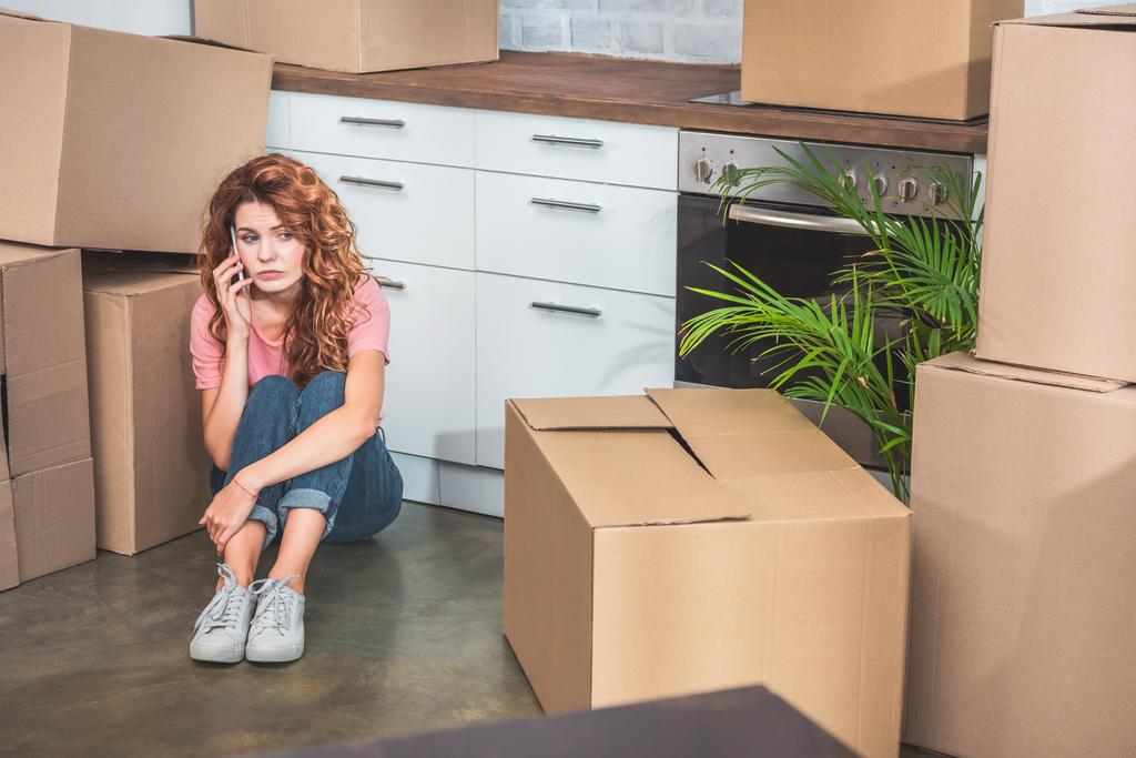 mujer triste en ropa casual sentada en el suelo cerca de cajas de cartón y hablando por teléfono inteligente en el nuevo hogar
 - Foto, Imagen