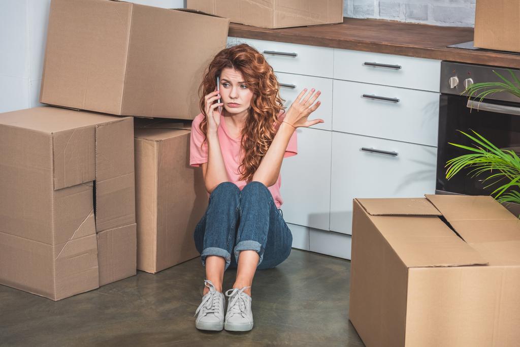 irritated woman with curly hair sitting on floor near cardboard boxes, talking by smartphone and gesturing at new kitchen - Photo, Image