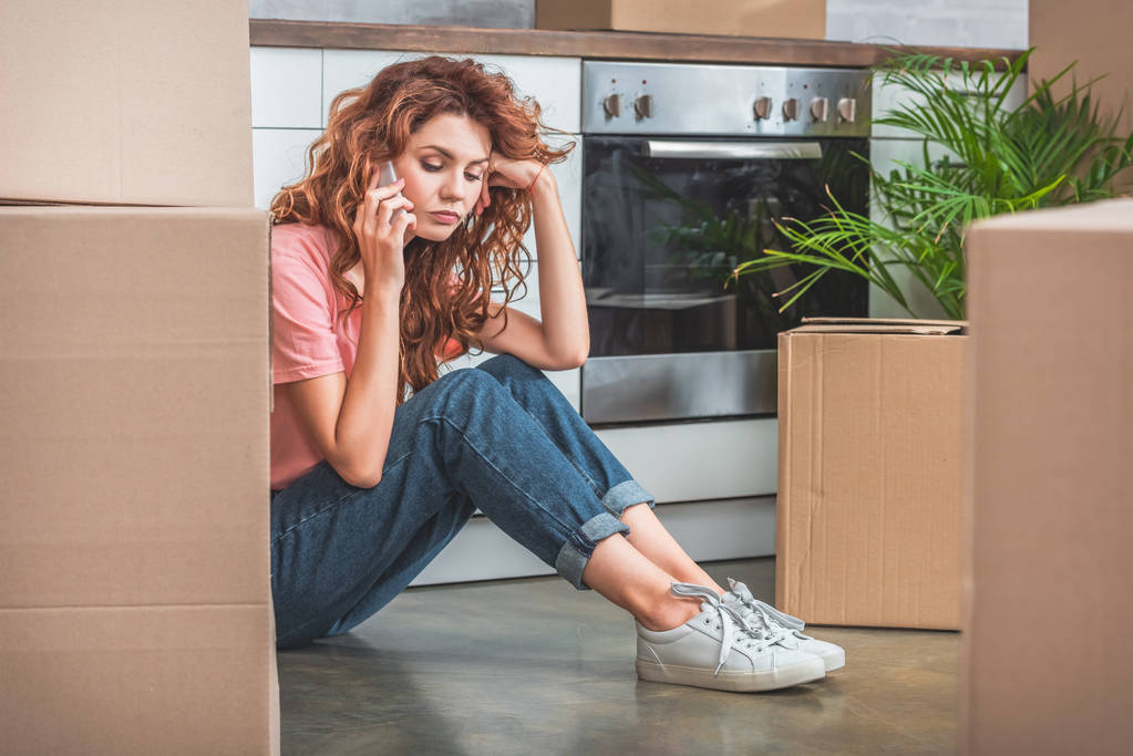 mujer triste con el pelo rizado sentado en el suelo cerca de cajas de cartón y hablando por teléfono inteligente en la nueva cocina
 - Foto, Imagen