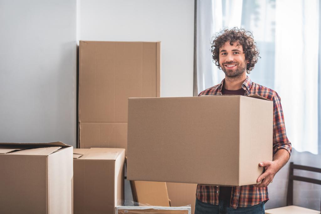 sonriente hombre guapo con pelo rizado sosteniendo caja de cartón en el nuevo hogar
 - Foto, imagen