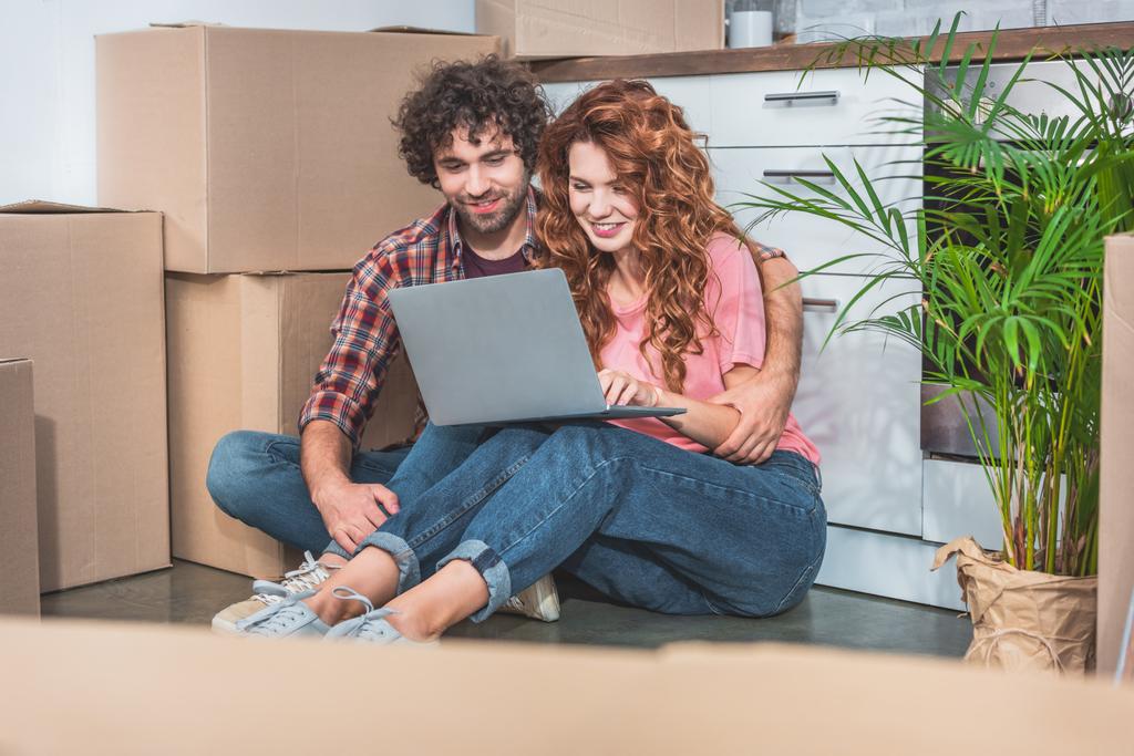 pareja abrazando y sentado con el ordenador portátil cerca de cajas de cartón en el suelo en la cocina nueva
 - Foto, imagen
