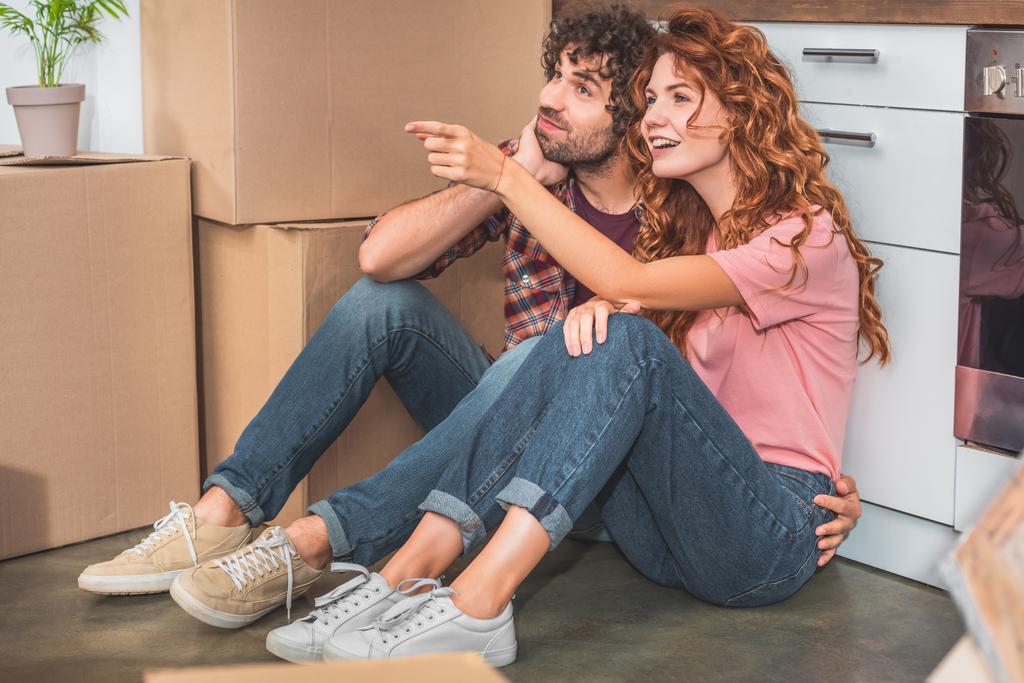 couple sitting on floor near cardboard boxes in new kitchen, girlfriend pointing on something - Photo, Image