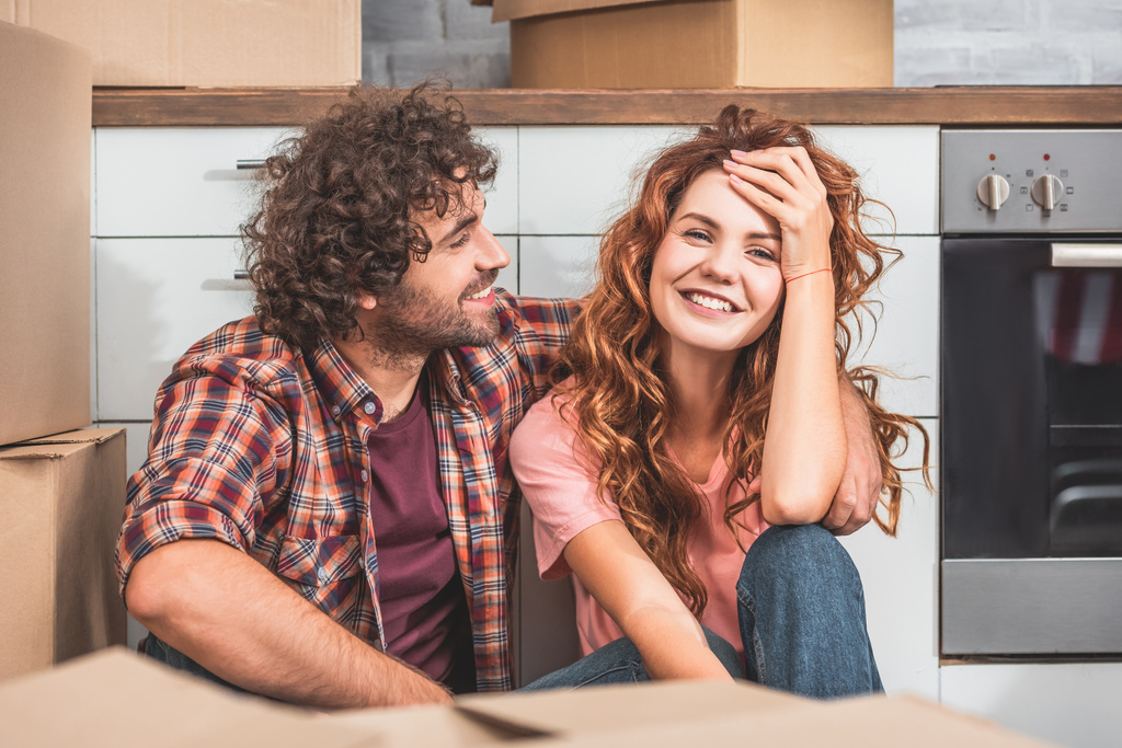 smiling couple sitting on floor near cardboard boxes in new kitchen - Photo, Image