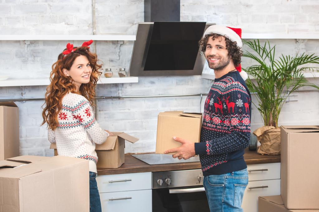 karton kutuları açma ve yeni evinde Noel yerini değiştirme sırasında kameraya gülümseyen mutlu genç çift - Fotoğraf, Görsel