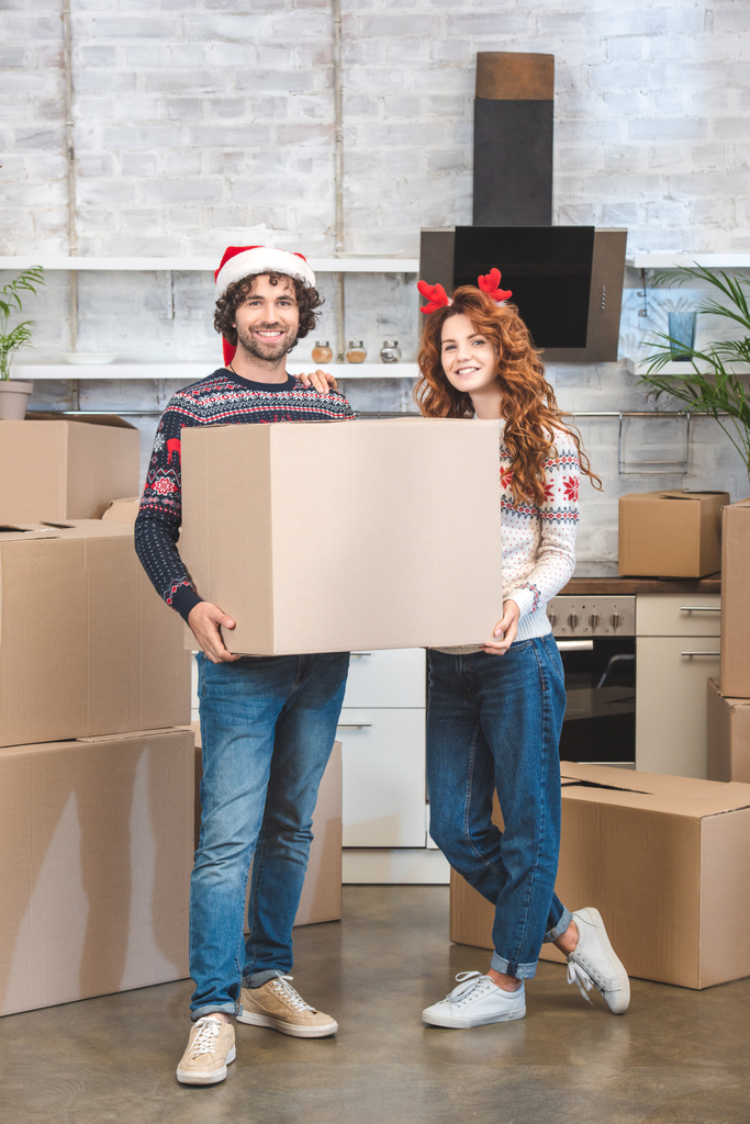 happy young couple holding cardboard box and smiling at camera while standing in new apartment at christmastime - Photo, Image