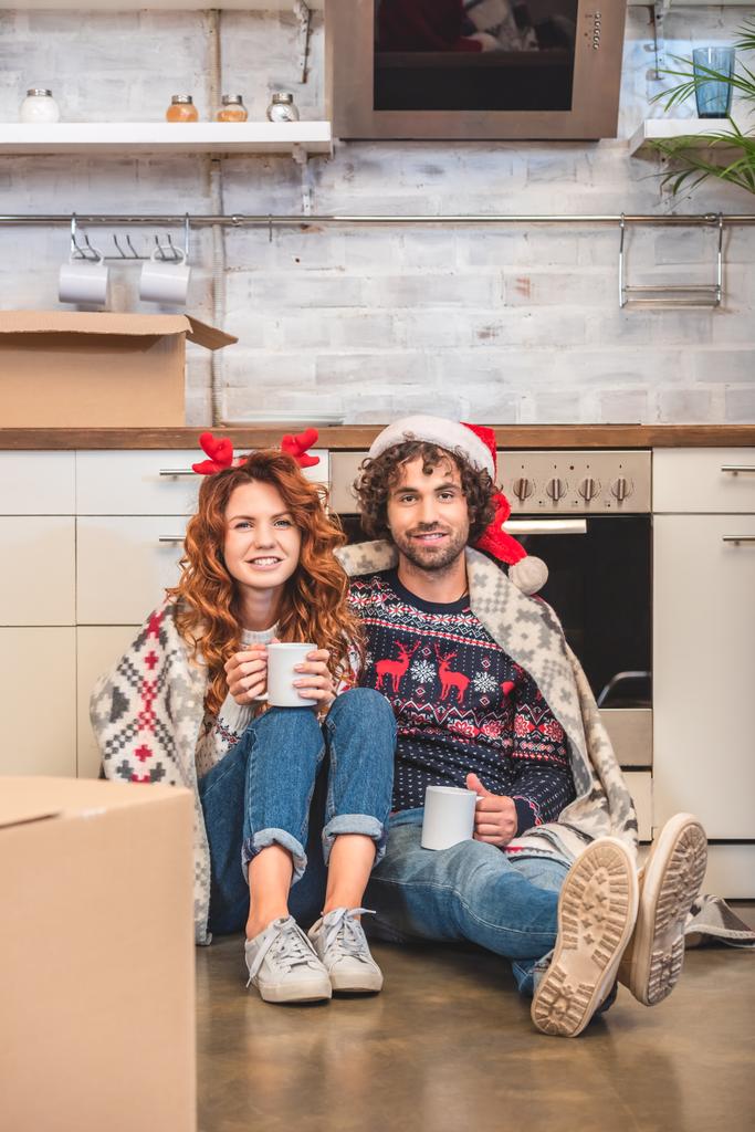 boldog fiatal pár holding kupa, és mosolyogva kamera ülve új lakás a christmastime - Fotó, kép