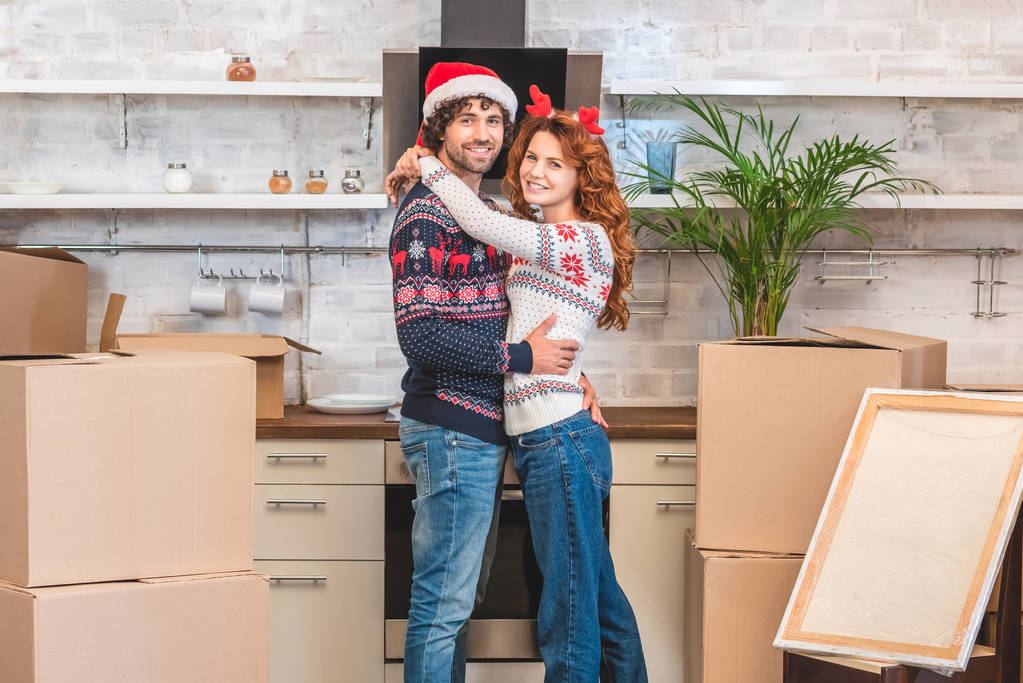 glückliches junges Paar umarmt und lächelt in die Kamera, während es zur Weihnachtszeit zwischen Pappkartons in seiner neuen Wohnung steht - Foto, Bild