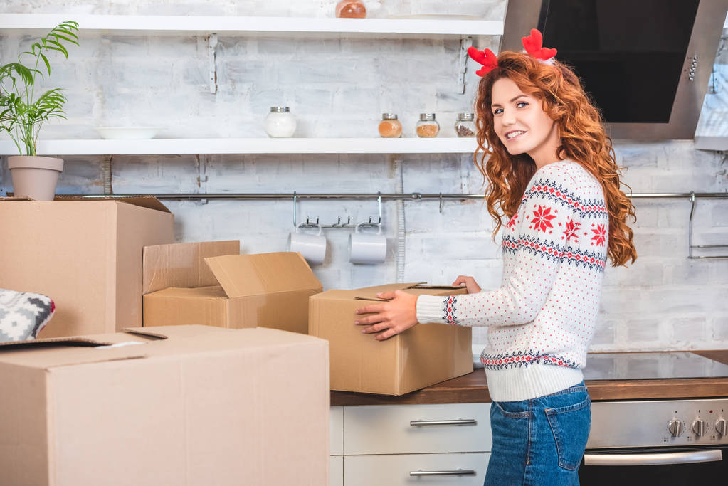 happy young woman in antlers headband unpacking cardboard boxes and smiling at camera during relocation - Photo, Image