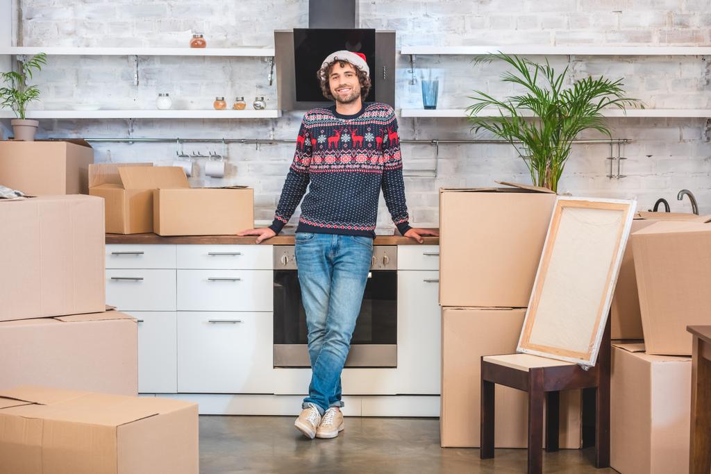 happy young man in santa hat smiling at camera while standing between cardboard boxes in new home - Photo, Image