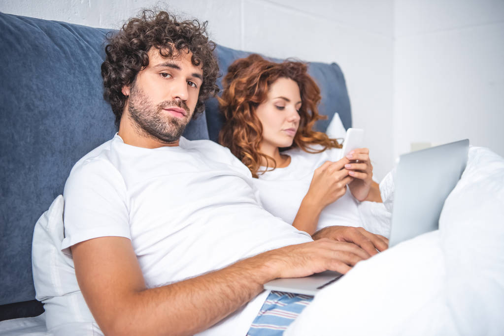 pareja joven usando el ordenador portátil y el teléfono inteligente en la cama
  - Foto, imagen