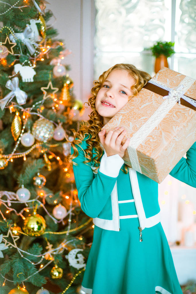 Menina bonito com cabelo louro encaracolado em casa perto de uma árvore de Natal com presentes e guirlandas e uma lareira decorada sentado em xadrez e travesseiros
 - Foto, Imagem