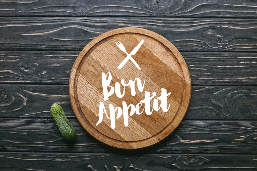 Cutting board with "bon appetit" lettering and cucumber on dark wooden table - Photo, Image