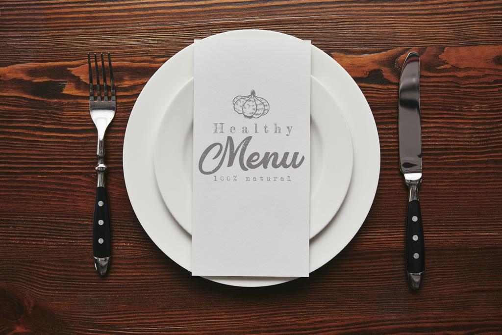 вид зверху карти з написом "здорове меню" на білих тарілках з виделкою і ножем на дерев'яному столі
  - Фото, зображення