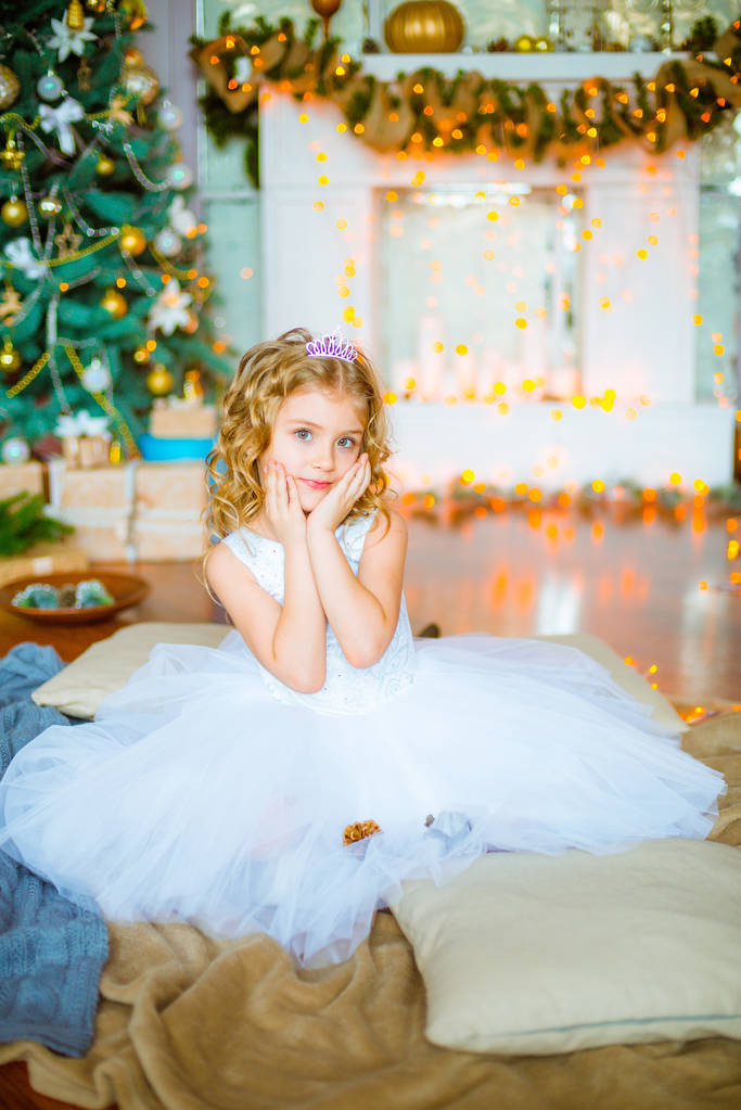 金髪巻き毛自宅ギフトと花輪と枕の上に座って装飾が施された暖炉とクリスマス ツリーの近くでかわいい女の子 - 写真・画像