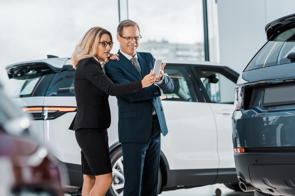 бизнесмен и деловая женщина со смартфоном выбирают новый автомобиль в выставочном зале
 - Фото, изображение