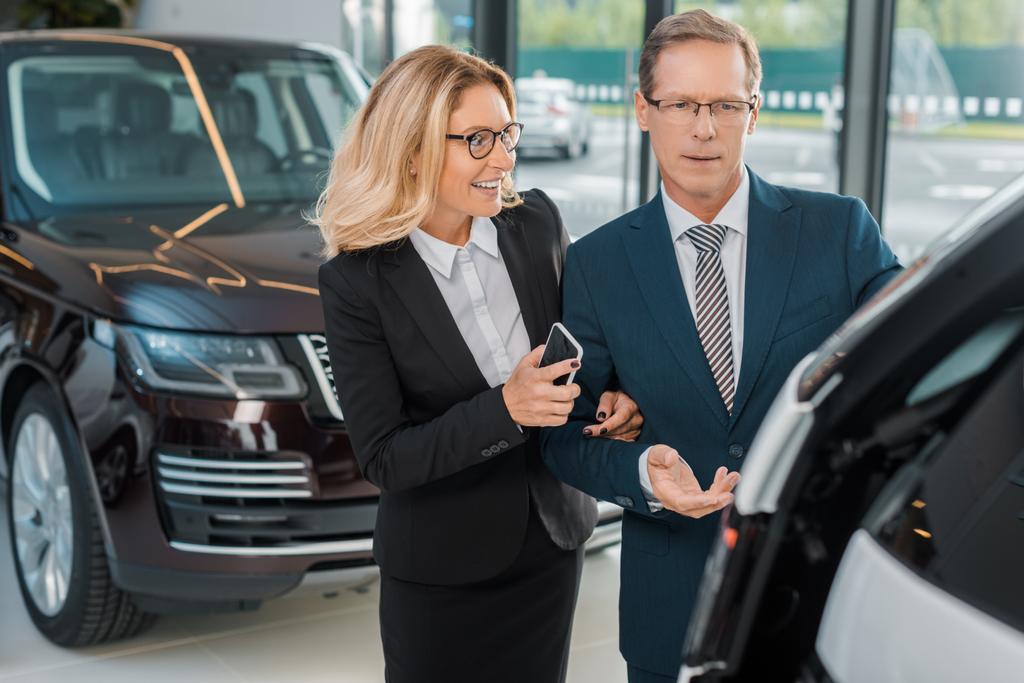 businessman and smiling businesswoman with smartphone choosing new car in dealership salon - Photo, Image