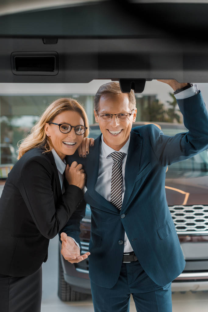 smiling couple in formal wear looking at new car in dealership salon - Photo, Image