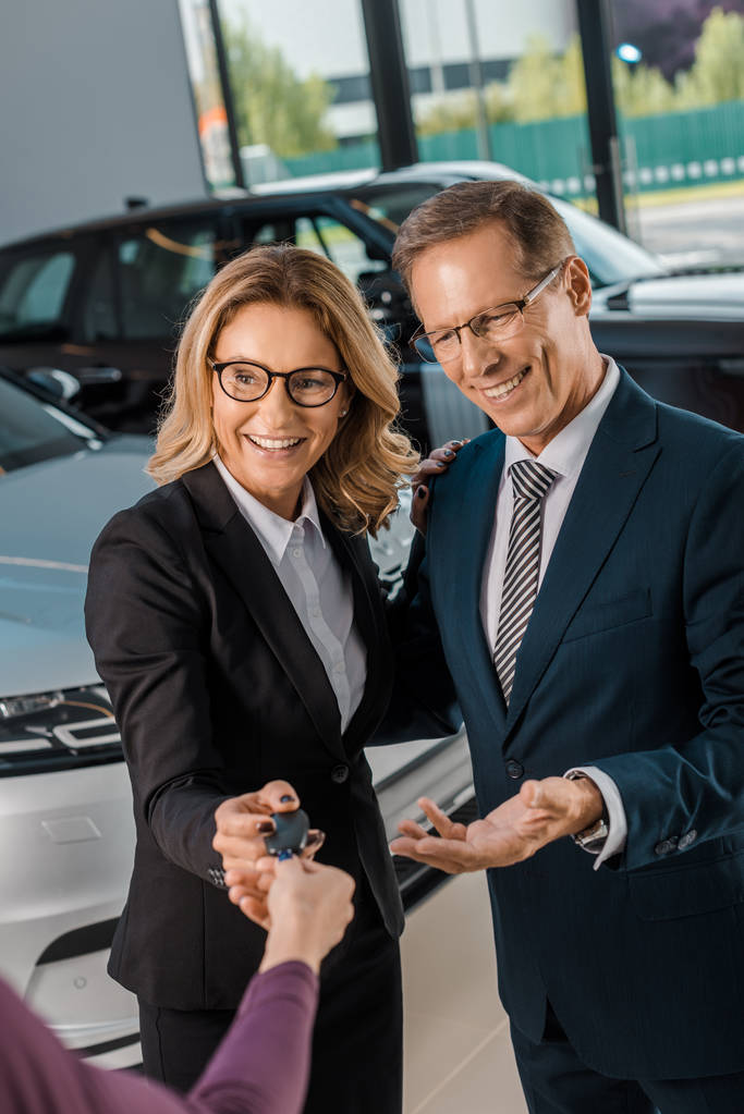 vista parcial de concesionario de coche femenino que da la llave del coche a la pareja de negocios sonriente en el desgaste formal en salón de concesionarios
 - Foto, Imagen
