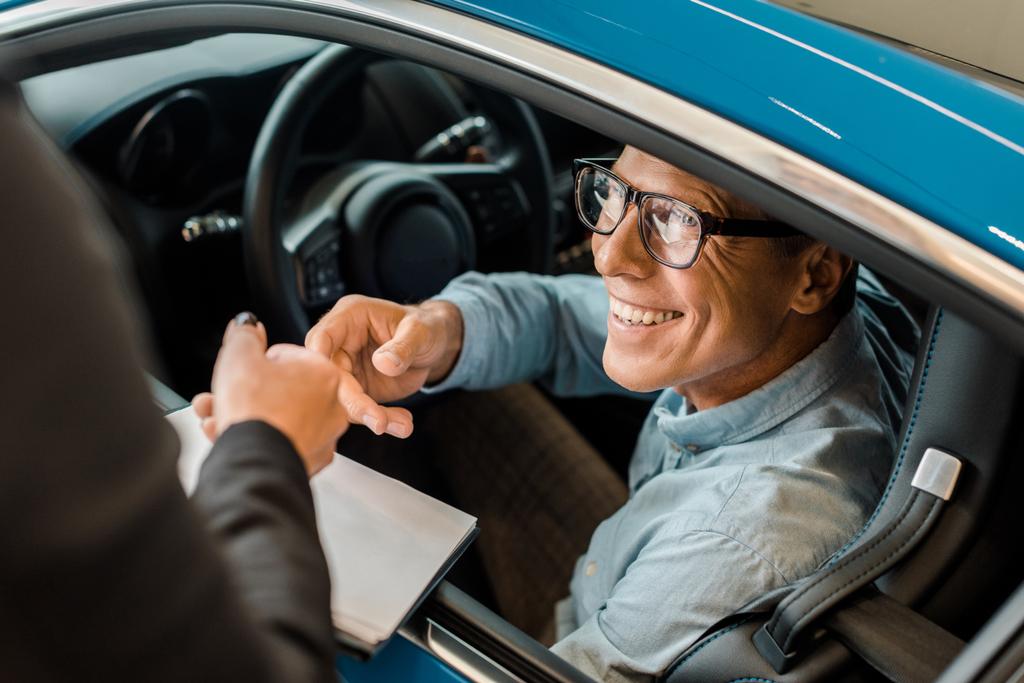 Schnappschuss einer Autohändlerin, die einem lächelnden erwachsenen Mann im Showroom den Händlervertrag überreicht - Foto, Bild