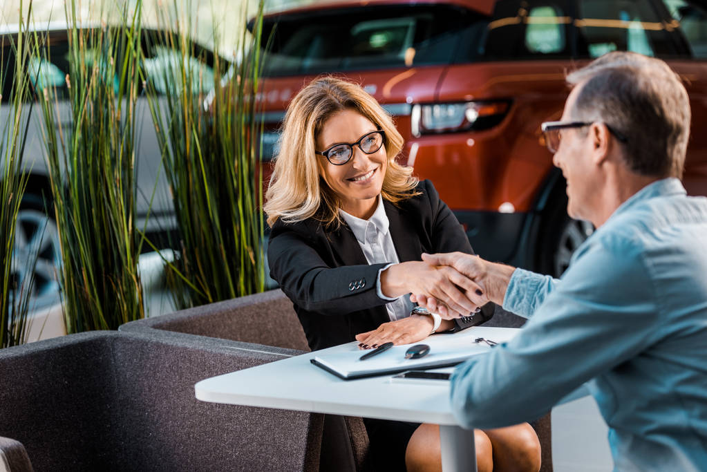 happy adult customer and female car dealer shaking hands in showroom - Photo, Image