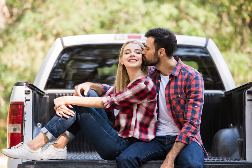 Mann küsst Wange einer glücklichen Freundin, während er auf Pickup-Truck sitzt   - Foto, Bild