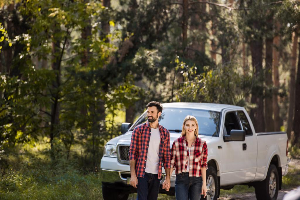 beautiful couple holding hands and standing near pickup truck in forest - Photo, Image