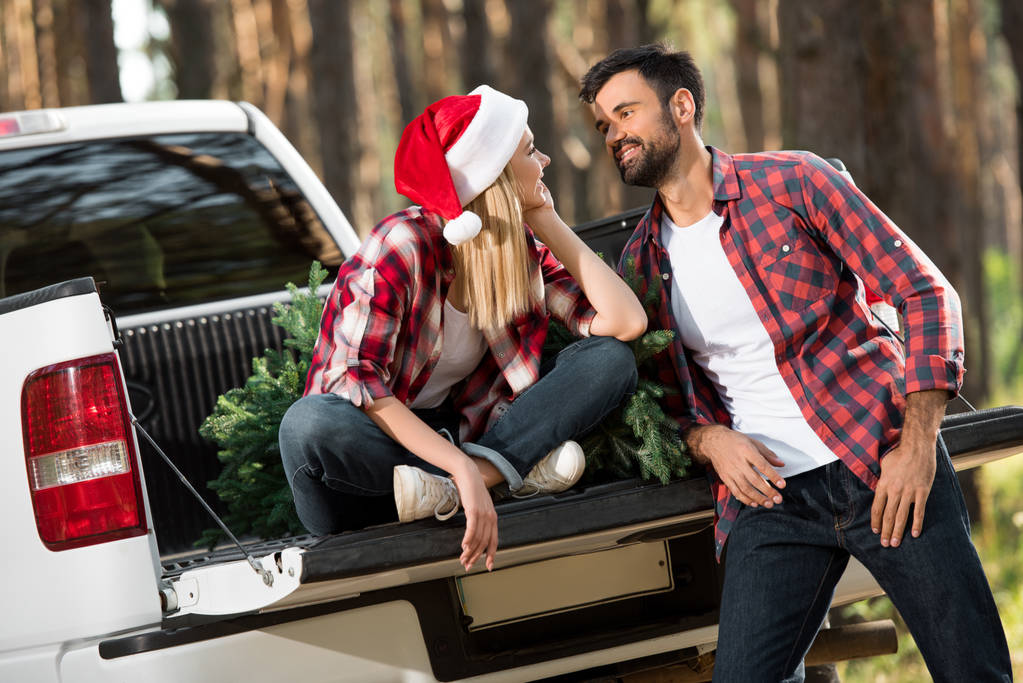 selective focus of woman in santa hat talking with boyfriend while sitting in car trunk with christmas tree in forest - Photo, Image
