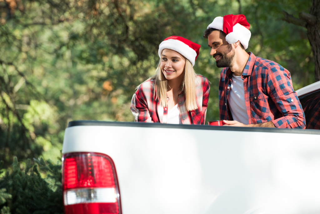 smiling young couple in santa hats with cups sitting in car trunk in forest  - Photo, Image