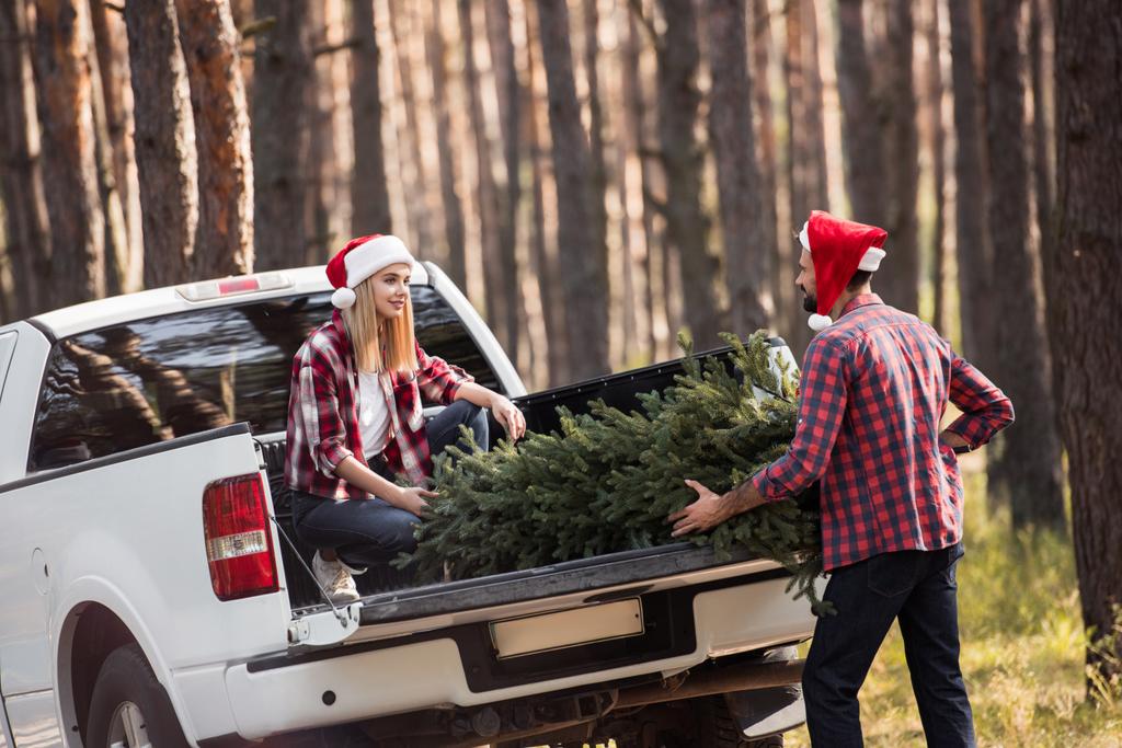 junges Paar in Weihnachtsmützen trägt Tannenbaum zu Weihnachten in Pickup-Truck im Wald - Foto, Bild