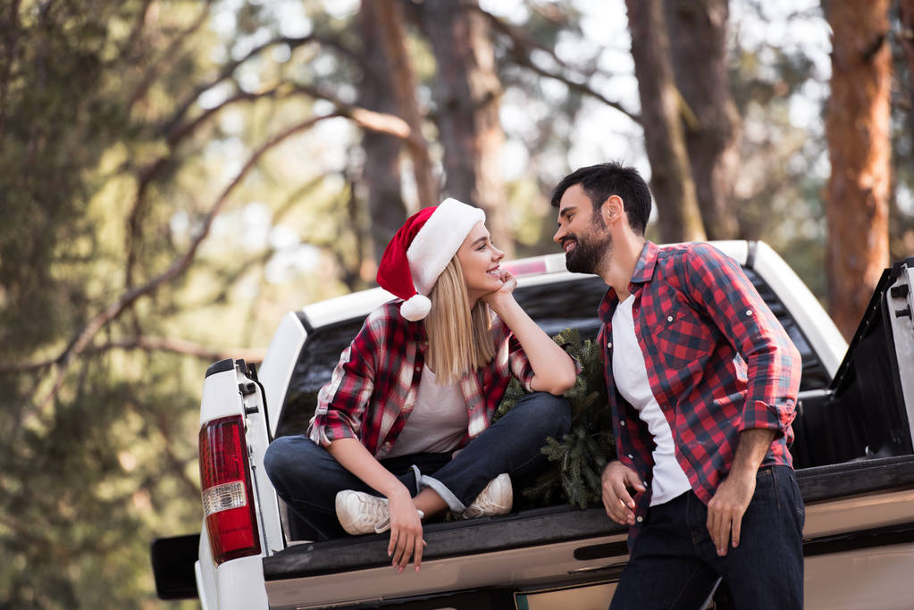 smiling couple in santa hats looking at each other and sitting on pickup truck with fir tree for christmas in forest - Photo, Image