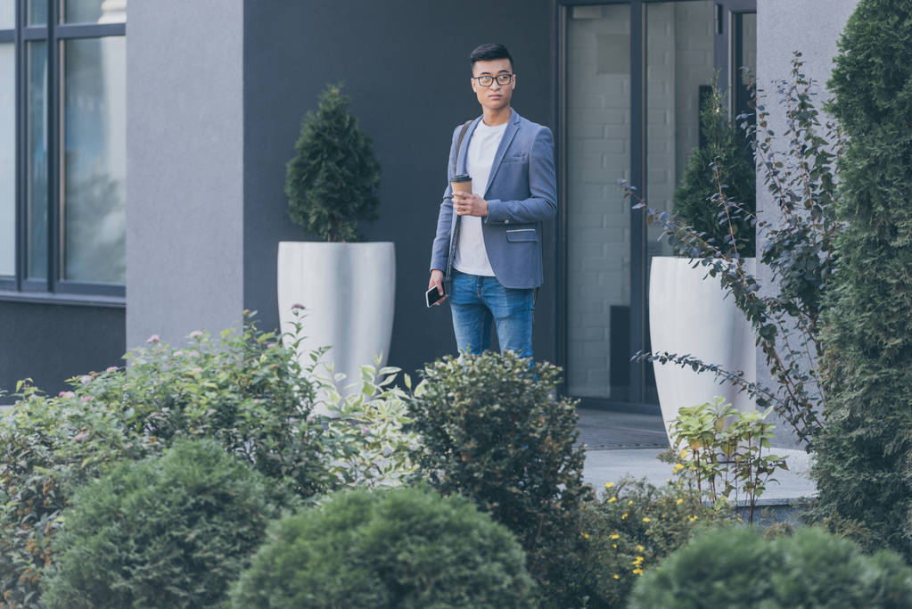 guapo asiático hombre sosteniendo café para ir y smartphone en la calle
 - Foto, imagen