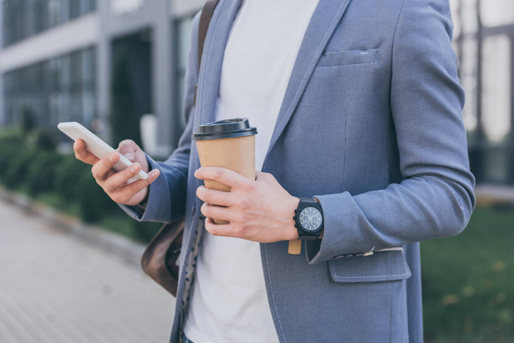 vista recortada del hombre en chaqueta gris que sostiene la taza de café desechable y el uso de teléfono inteligente
  - Foto, Imagen