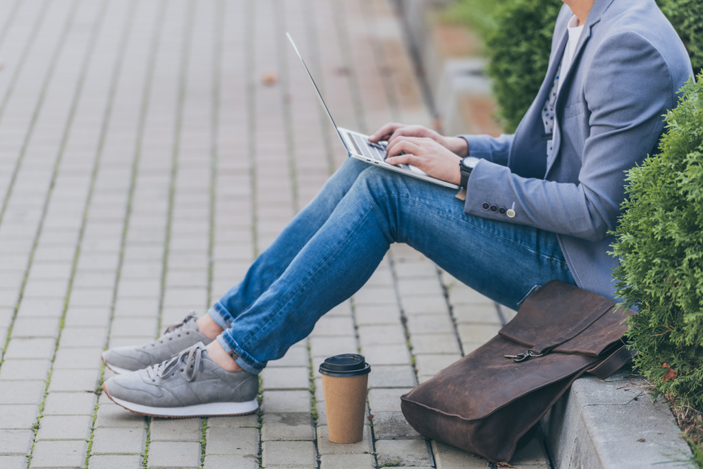 Ausgeschnittene Ansicht eines Freiberuflers, der am Laptop arbeitet, während er mit Ledertasche und Coffee to go auf dem Bürgersteig sitzt - Foto, Bild