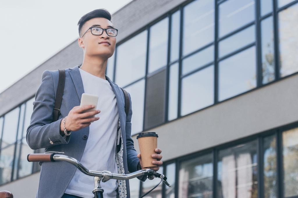 stylish confident asian man holding disposable cup of coffee and smartphone near bike in city - Photo, Image