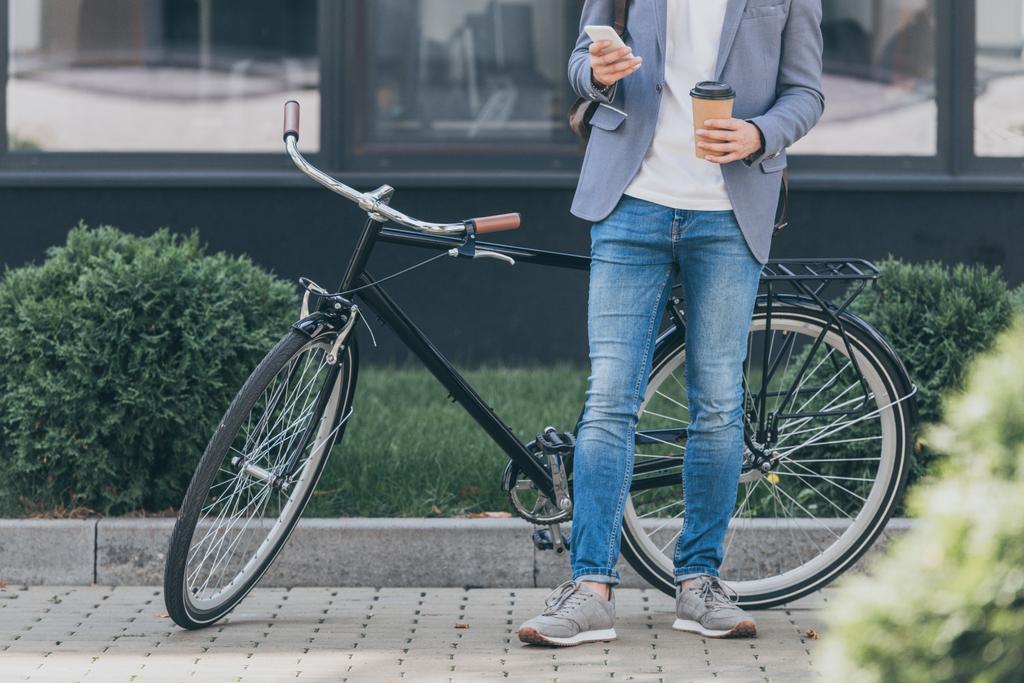 oříznutý pohled stylové muže s kávou jít pomocí smartphone a stojící poblíž kolo - Fotografie, Obrázek