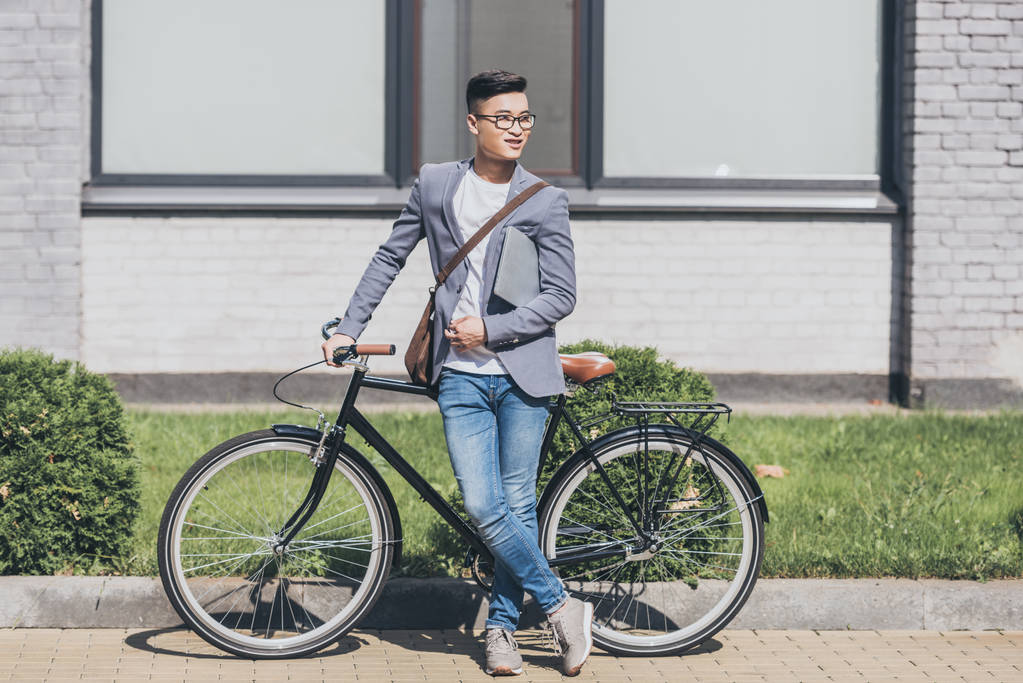 smiling asian man in trendy jacket holding laptop and standing at bicycle - Photo, Image