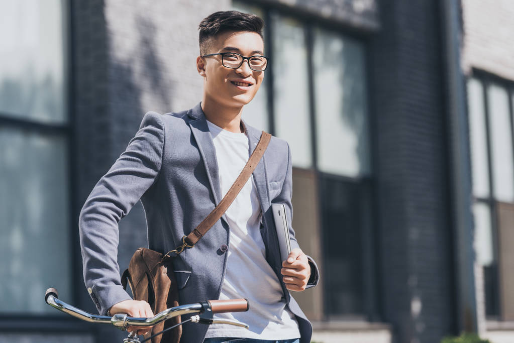 uomo asiatico sorridente con computer portatile e borsa in pelle in piedi con bicicletta
 - Foto, immagini