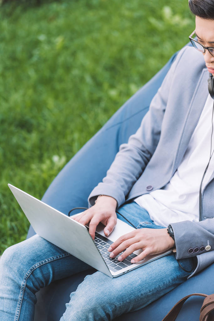 Freelancer asiático trabalhando no laptop enquanto sentado na cadeira de saco de feijão no gramado
 - Foto, Imagem