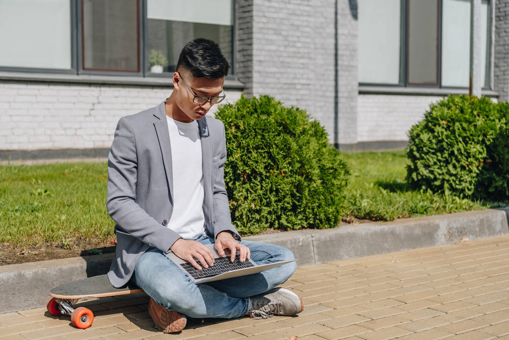 Ασιατική άνθρωπο πληκτρολογώντας στον φορητό υπολογιστή ενώ κάθεστε στον longboard στην οδό - Φωτογραφία, εικόνα
