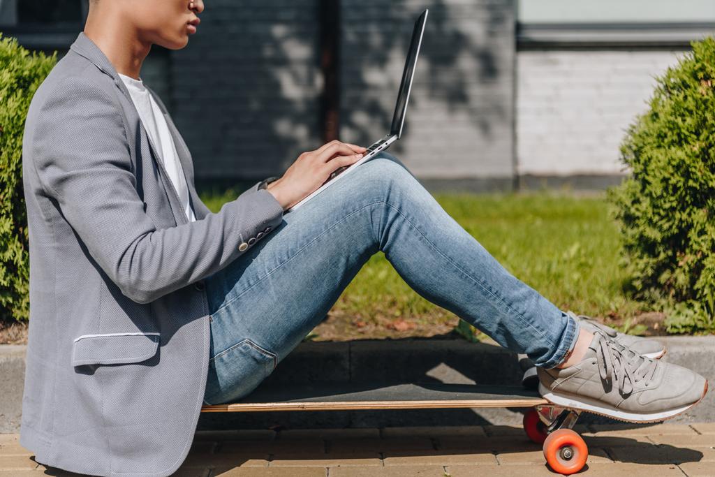 partial view of teleworker using laptop while sitting on longboard on street - Photo, Image