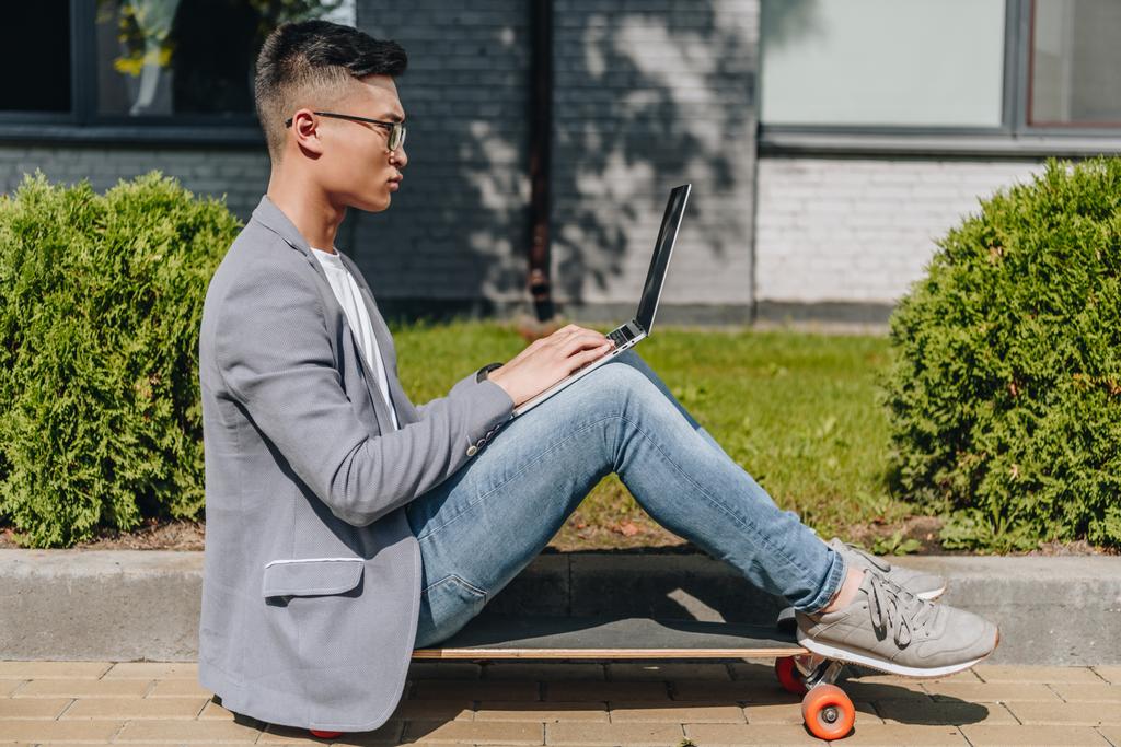 вид збоку азіатського фрілансера, який використовує ноутбук, сидячи на дошці на вулиці
 - Фото, зображення