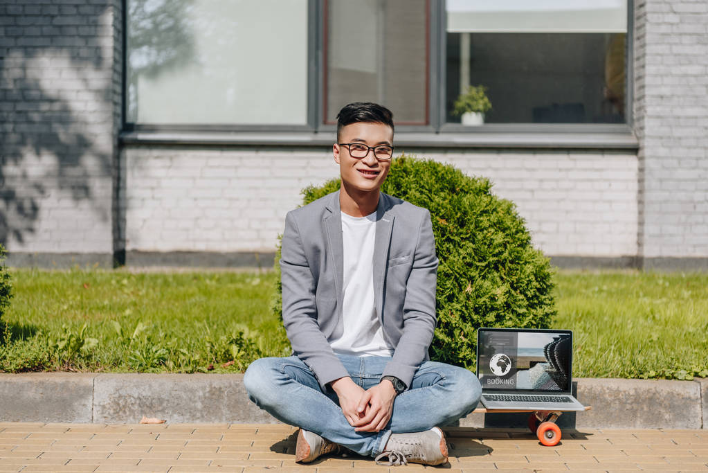 lächelnder asiatischer Mann sitzt auf Longboard mit Laptop mit Buchungswebseite auf dem Bildschirm auf der Straße - Foto, Bild