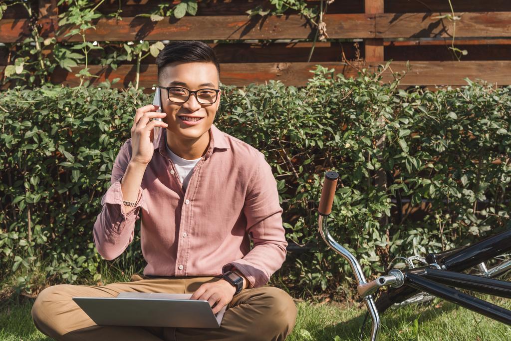 smiling asian man talking on smartphone while remote working on laptop in park - Photo, Image
