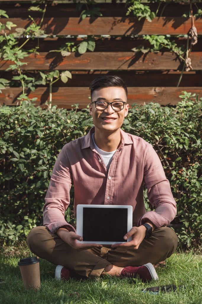 sonriente asiático hombre sentado en verde hierba y mostrando tableta con pantalla en blanco en las manos en parque
 - Foto, Imagen
