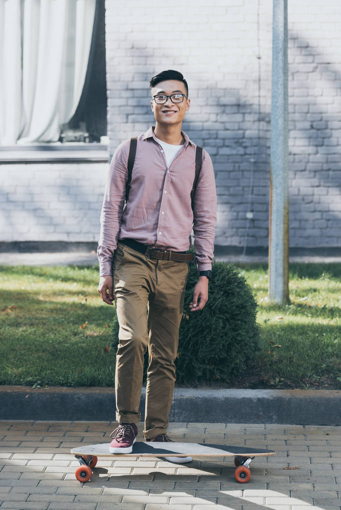 Lächelnder asiatischer Mann mit Rucksack und Longboard blickt auf der Straße in die Kamera - Foto, Bild