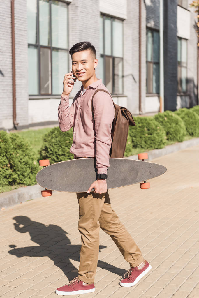 vista lateral de sorrir asiático homem com longboard tendo conversa no smartphone na rua
 - Foto, Imagem